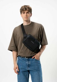 Borsa a tracolla nera realizzata in materiale resistente; presenta una forma rettangolare, una tasca con zip e una tracolla regolabile. Uomo che indossa una camicia marrone.