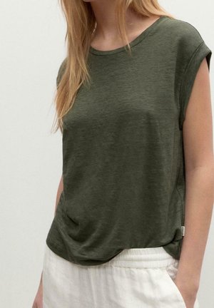 Frau trägt ein ärmelloses dunkelgrünes Oberteil, das in lockere weiße Hose gesteckt ist, steht mit einer Hand in der Tasche vor einfarbigem Hintergrund.