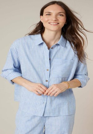 Vrouw met lang bruin haar die lacht, gekleed in een lichtblauw-wit gestreept overhemd met knopen en bijpassende broek, handen samengevouwen ter hoogte van de taille.