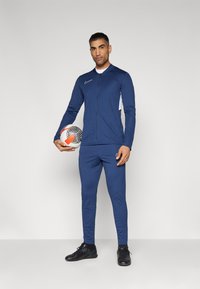 Marineblauw sporttrack suit van lichtgewicht stof, met een ritsjack met witte accenten. Het model houdt een voetbal vast.