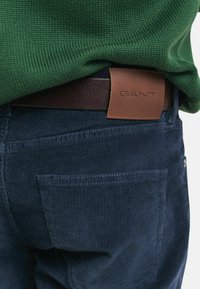 Marine corduroy broeken met een gestructureerde stof; bruine leren GANT logo patch op de tailleband; groene gebreide trui gedeeltelijk zichtbaar.