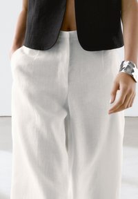 Weißes Leinen weit geschnittene Hose mit glatter Textur, kombiniert mit einem schwarzen Cropped-Top. Ein silbernes Armreif ist am Handgelenk.
