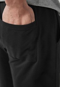 Shorts negri din bumbac cu o buzunar lateral, având o textură netedă și linii curate. Mână plasată în buzunar.