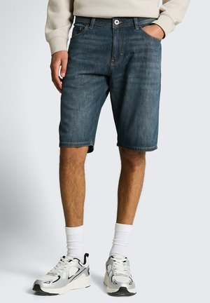 Mannequin masculin portant un short en jean foncé de longueur moyenne, des chaussettes blanches, des baskets blanches et noires, et un sweat-shirt beige avec les mains partiellement dans les poches.