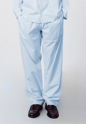 Mężczyzna stojący z rękami w kieszeniach, ubrany w jasnoniebieskie spodnie i ciemnobrązowe eleganckie buty na neutralnym tle.