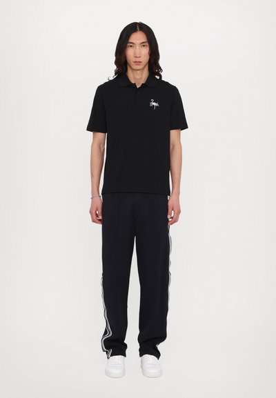 Black polo shirt with short sleeves and a flamingo logo, paired with navy trousers featuring white side stripes and white sneakers.