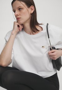 White oversized cotton t-shirt with a round neckline and short sleeves. Notable small embroidered design on the chest. Black leggings and handbag.