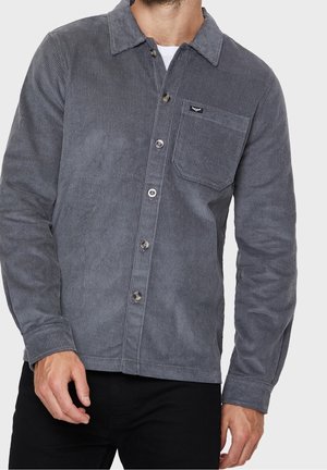 Mężczyzna ubrany w ciemnoszarą koszulę na guziki z mankietami z sztruksu i czarne spodnie, stojący na jednolitym tle.