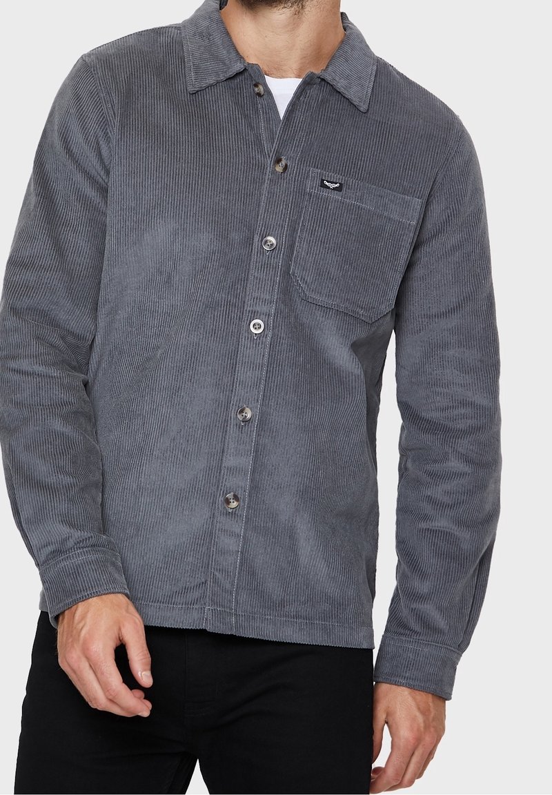 Homme portant une chemise en velours côtelé gris foncé à boutons avec poche poitrine et un pantalon noir, debout devant un fond uni.