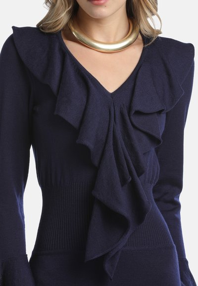 Mornarsko modra jopica z nabranim detajlom na izrezu, rebrasto teksturo na rokavih in telesu, dopolnjena z zlatim statement nakitom.