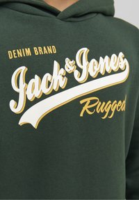 Zelená mikina s kapucí s bílým a žlutým nápisem "Jack & Jones Rugged" a zakřiveným bílým akcentem. Měkký, bavlněný materiál s volným střihem.