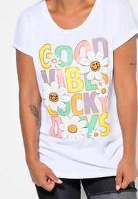 T-shirt blanc avec une impression textuelle colorée disant "BONNES VIBES JOURS CHANCEUX", embellie de motifs de marguerites et d'une texture lisse.