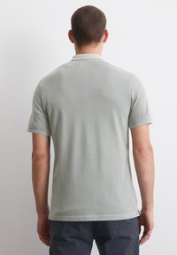 Mężczyzna widziany z tyłu, ubrany w jasnoszary koszulowy T-shirt z krótkim rękawem i ciemne spodnie na jednolitym białym tle.