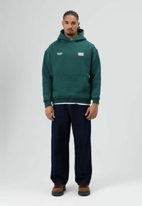Groene hoodie met een voorzak en logo's, gecombineerd met losse donkere jeans en bruine schoenen. De stof lijkt glad en dik.