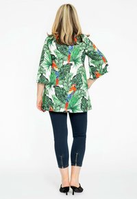 Tunique florale avec des feuilles vertes et des perroquets colorés, associée à un pantalon ajusté bleu foncé avec des accents zippés à la cheville, portée avec des talons noirs.