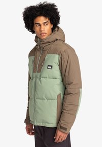 Puffjacka i brunt och mintgrönt, med huva, dragkedjeförsedda främre fickor och en logotyppatch på vänster bröst, med ett strukturerat tyg.