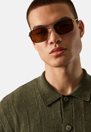 Ung mand iført rektangulære solbriller med brune nuancer og en grøn tekstureret polo-shirt mod en ensfarvet baggrund.
