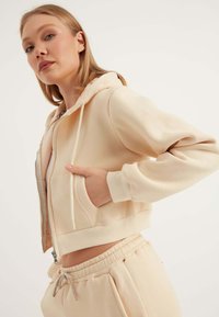 Felpa corta con zip in morbido tessuto beige, dotata di cappuccio, tasche frontali e polsini a costine. Abbinata a jogger beige coordinati.