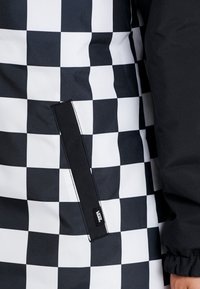 Svart och vit rutigt jackficka med svart kant och Vans-logotagg, bredvid en svart ärm med elastisk mudd.