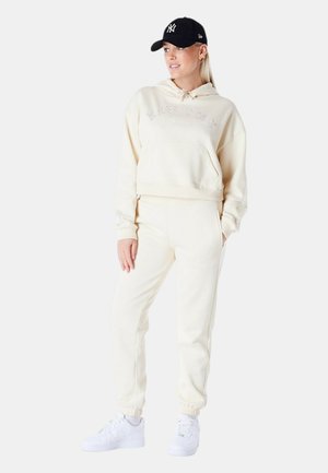 Mujer con sudadera con capucha crema y pantalones deportivos, zapatillas blancas y gorra negra de los New York Yankees, de pie con una mano en el bolsillo.