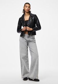 Must musta biki jakk mustast nahast, millel on sobiv disain, kombineerituna laia säärega heledate hallide teksadega. Mudel seisab ühtlase valge tausta ees.