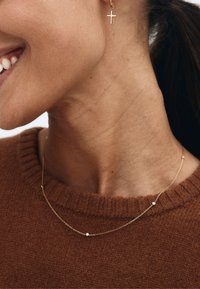 Zlatý náhrdelník s jemným dizajnom, zdobený malými korálkami. Modelka má na sebe hnedý sveter a zlatú náušnicu v tvare kríža. Hladká textúra.