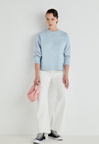 Lichtblauwe sweatshirt met ronde hals en lange mouwen, gecombineerd met witte wijde broek en zwarte sneakers. Houdt een roze tas vast.