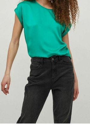 Frau trägt eine leuchtend grüne kurzärmelige Bluse, die in eine hoch taillierte, ausgewaschene schwarze Jeans gesteckt ist, und steht mit entspannten Armen an den Seiten.