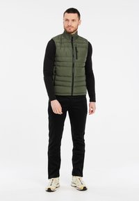 Olivengrüne gepolsterte Weste mit hohem Kragen, Frontreißverschluss und seitlichen Taschen. Getragen über einem schwarzen Langarmshirt und kombiniert mit schwarzen Hosen.