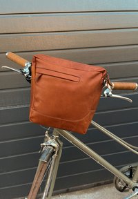 Bolsa de cuero marrón sujeta al manillar de una bicicleta. Cuenta con un cierre de cremallera, un bolsillo frontal y una textura suave. La bolsa tiene forma cuadrada.