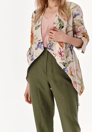 Femme portant une veste beige à fleurs ouverte sur le devant, un chemisier rose et un pantalon taille haute vert olive, une main tenant le devant de la veste.
