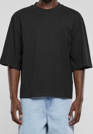 Osoba ubrana w luźny czarny t-shirt z krótkim rękawem i jasnoniebieskie dżinsy na jednolitym szarym tle.