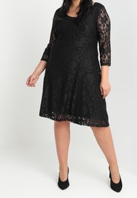 Svart spetsklänning med utsvängd kjol, trekvartsärmar och rundad halsringning. Har intrikata blommönster och en texturerad yta.