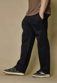 Personne portant un pantalon noir et des baskets noir et gris, se tenant avec les mains dans les poches devant un fond beige.