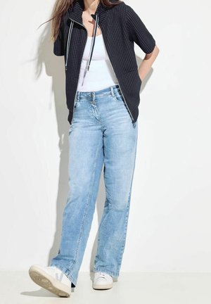 Femme portant un jean bleu clair, des baskets blanches, un débardeur blanc et une veste noire zippée à manches courtes, debout contre un mur blanc.