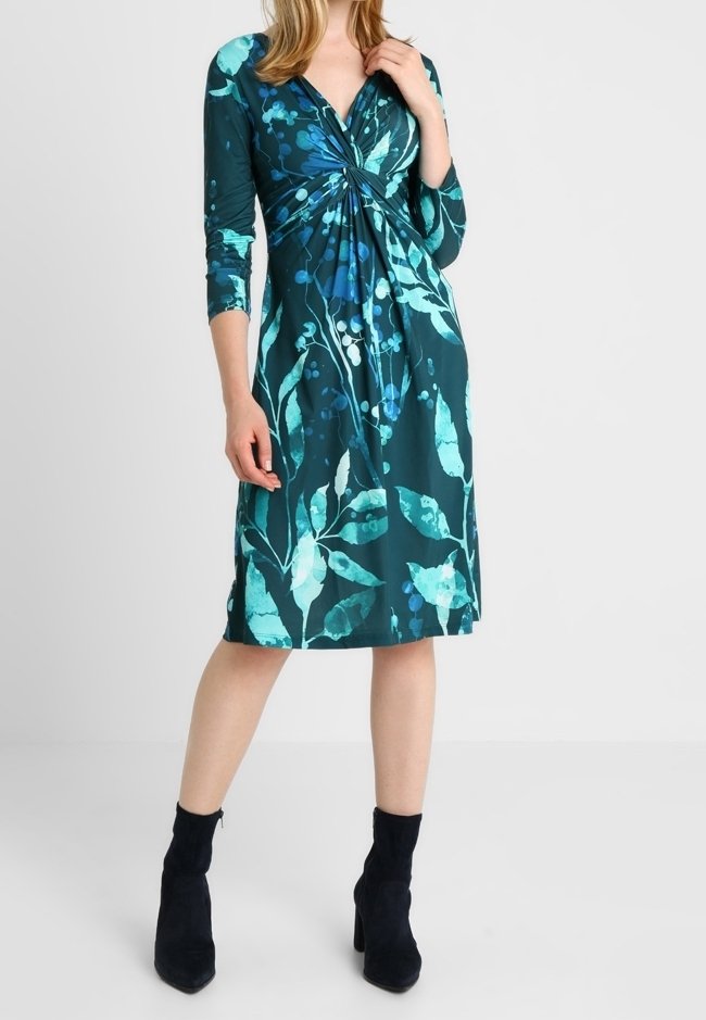Femme portant une robe fleurie bleu canard et bleu longueur genou avec manches trois-quarts et bottines noires, debout devant un fond uni.