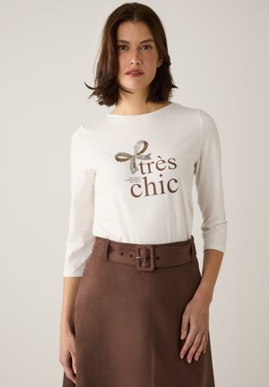 Weißes Langarmhemd mit "très chic" in Braun bedruckt und einem dekorativen Banddesign; kombiniert mit einem braunen Rock mit breitem Gürtel.