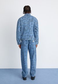 Jeansjacke und Hose in Hellblau mit einem subtilen Tarnmuster. Die Jacke hat seitliche Taschen, eine lockere Passform und einen spitzen Kragen. Schwarze Schuhe.