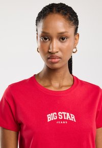 Sarkana kokvilnas T-kreklu ar īsām piedurknēm, kurā ir balts uzraksts "BIG STAR JEANS". Apaļa kakla izgriezums, gluda tekstūra un pieguļošs griezums.