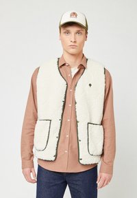Gilet in pile bianco con rifiniture verdi, dotato di due tasche frontali, indossato sopra una camicia a quadri marrone chiaro e jeans blu. Cappellino bianco con logo.