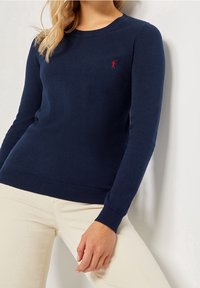 Námořnická modrá pletená mikina s dlouhými rukávy, žebrovanými manžetami a spodním lemem, s malým červeným logem na hrudi. Měkká textura a přiléhavý střih.