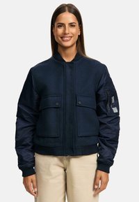 Navy bomber jacket with two front pockets, ribbed cuffs, and a smooth texture. Features a patch on the left sleeve with reflective detail.