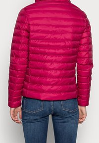 Istutettu, kirkkaan pinkki toppatakki, jossa vaakasuorat tikatut viivat, pystykaulus ja joustavat hihansuut, yhdistetty tummansinisiin farkkuihin.