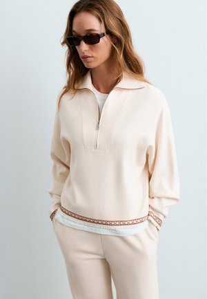 Woman wearing beige zip-up sweater and matching pants with hands in pockets and dark rectangular sunglasses against plain background.