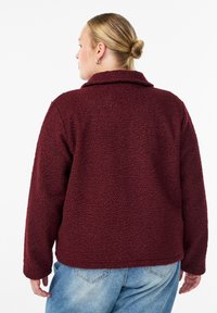 Veste en polaire bordeaux avec une finition texturée, un large col et des manches longues ; portée avec un jean bleu clair.