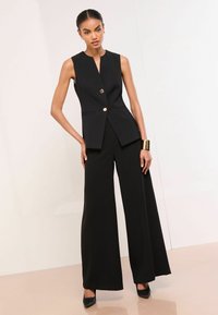 Schwarz ärmellose Weste mit V-Ausschnitt, Ein-Knopf-Verschluss und zwei Seitentaschen; kombiniert mit weit geschnittenen schwarzen Hosen und spitzen Schuhen.