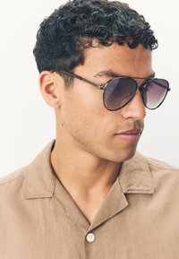 Naočale za sunce s tamnim gradijentnim staklima i tankim crnim metalnim okvirom. Nositelj nosi svijetlosmeđu košulju s ovratnikom i gumbom.