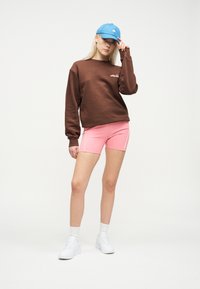 Νεαρή γυναίκα με καφέ φούτερ, ροζ σορτς, μπλε καπέλο, λευκές κάλτσες και λευκά αθλητικά παπούτσια, στέκεται μπροστά σε λευκό φόντο.