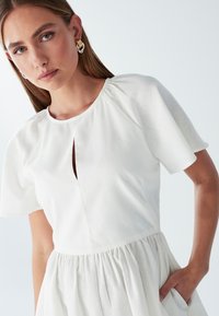 Biele šaty s krátkymi rukávmi, s okrúhlym výstrihom a výrezom v tvare kľúčovej dierky vpredu. Látka je hladká a sukňa je skladaná.