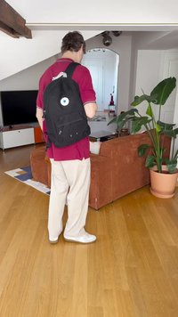 Μαύρο σακίδιο πλάτης με κυκλικό λογότυπο, πολλές τσέπες και φερμουάρ. Φοριέται από ένα άτομο με κόκκινο πουκάμισο και μπεζ παντελόνι, σε φωτεινό εσωτερικό χώρο.